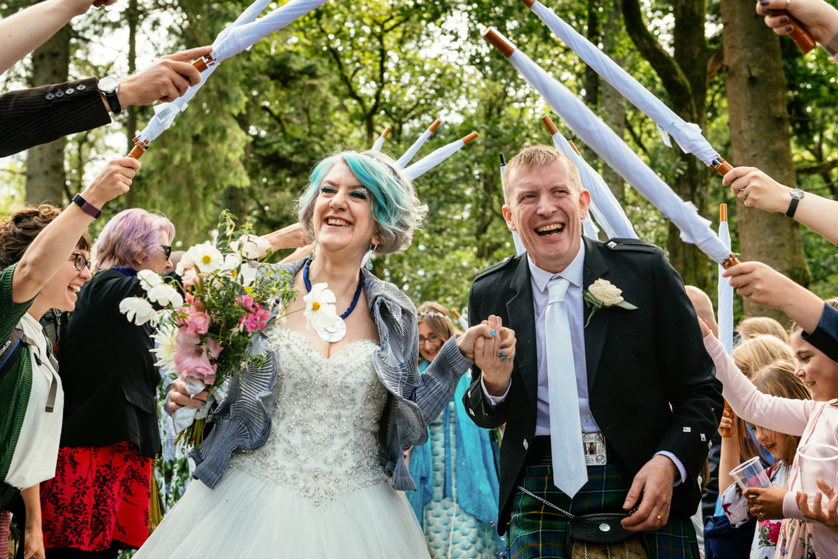 Couple walking through an archway formed by guests holding matching white umbrellas after their wedding ceremony in South Wales, captured by a wedding photographer