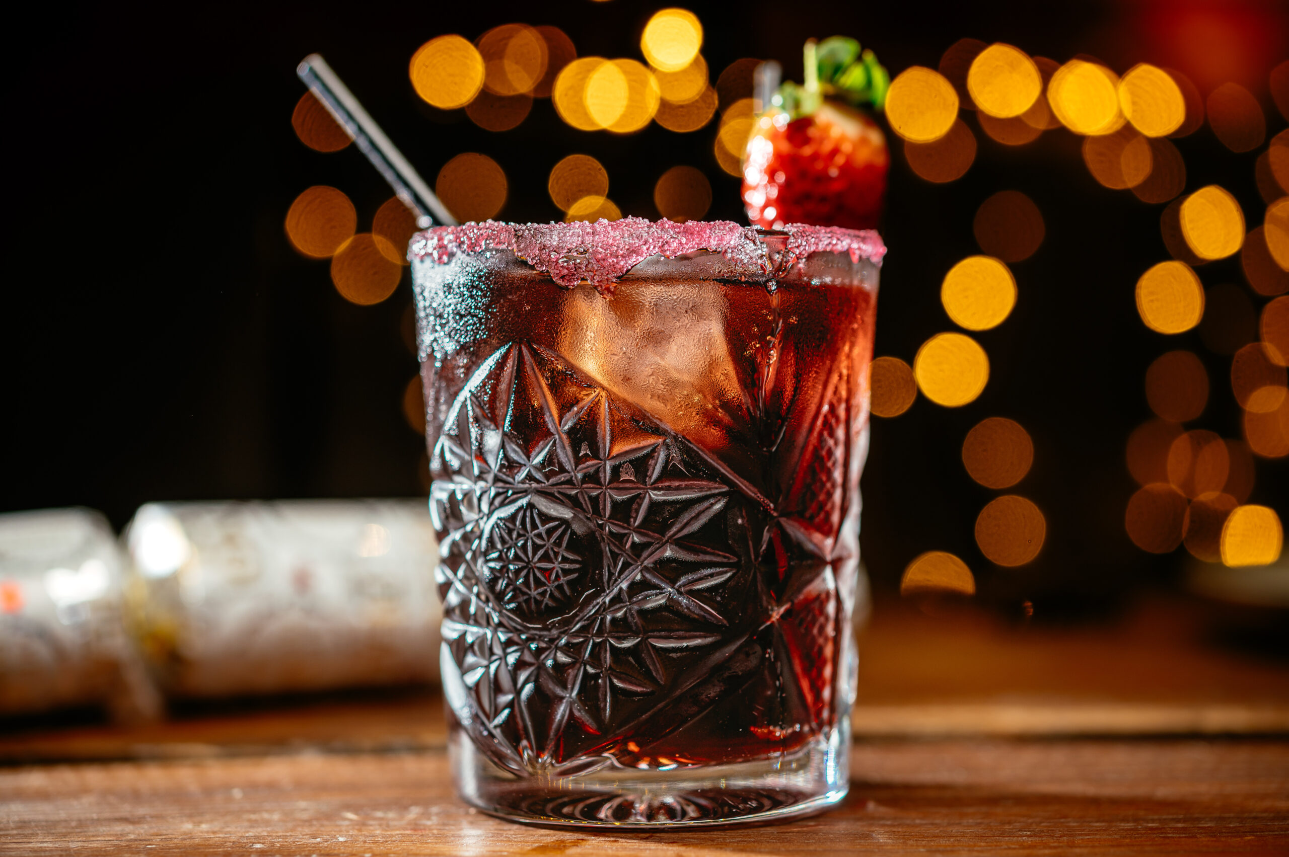 cocktail in a cut glass with brown liquid inside and pick sugar on the rim with a straw